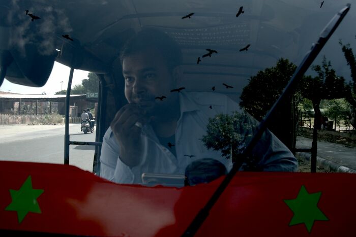 Man driving an auto-rickshaw, seen through a windshield with reflections, highlighting street photography awards.