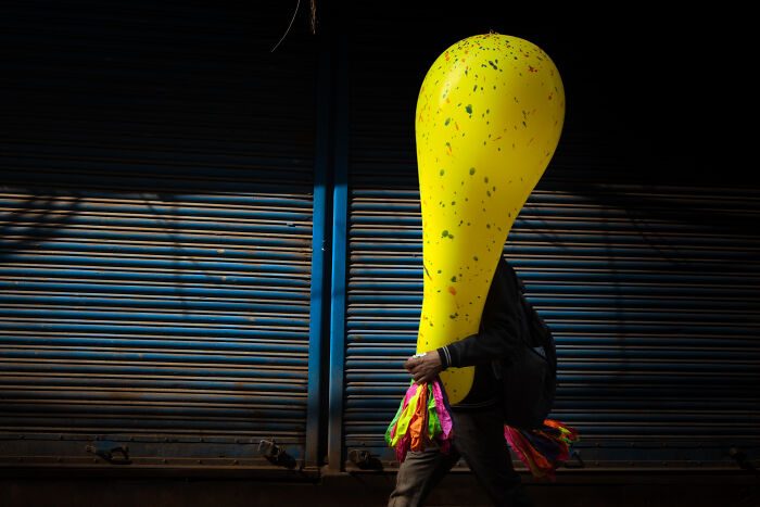 Man carrying a giant yellow balloon walking past blue shutters, capturing an authentic moment in street photography.