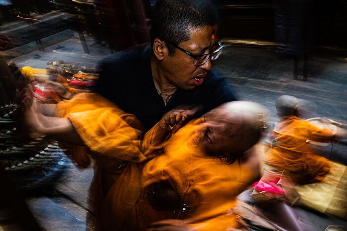 A blurred street photography image of a man in glasses carrying someone in an orange robe, capturing an authentic moment of life.