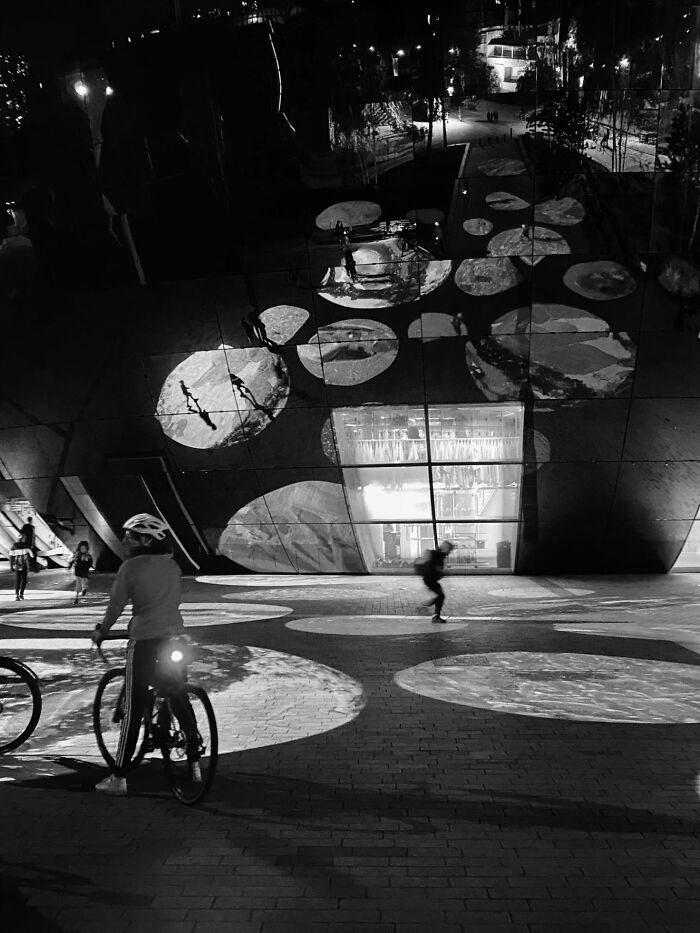 Nighttime street photography of a cyclist and pedestrians passing a building with reflective, illuminated patterns.