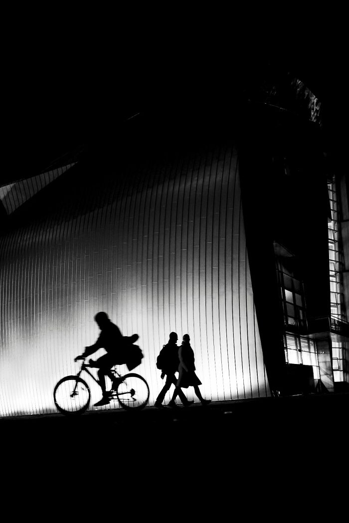Silhouettes of a person cycling and two pedestrians walking against a bright, textured wall at night, showcasing street photography.
