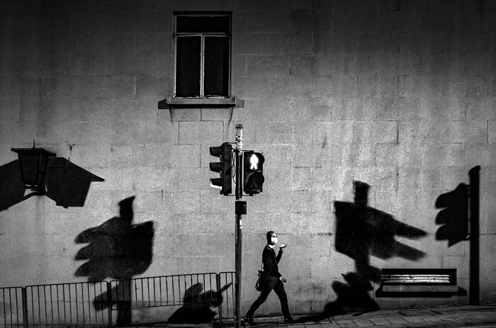 Pure street photography capturing a masked person walking past a traffic light with strong shadows.