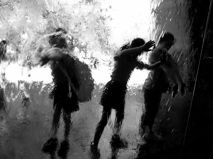 Pure street photography capturing blurred figures behind a textured glass or water wall, creating authentic moments.
