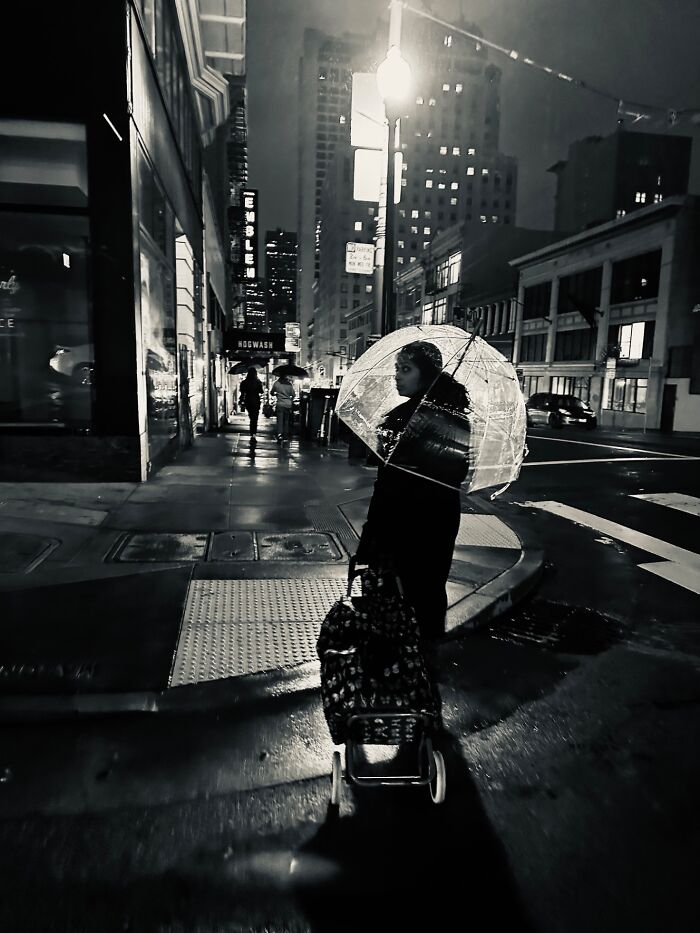 Woman with clear umbrella and shopping cart on a rainy city street at night.