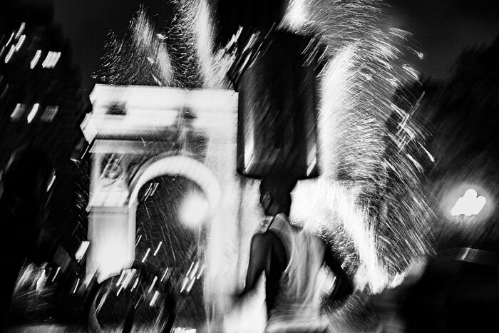 Blurred figure with a box on their head, amidst Washington Square Arch and fireworks.