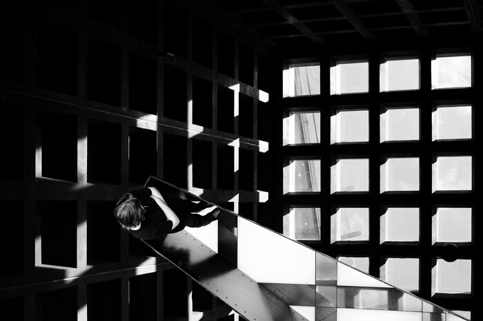 A person sitting on a checkered ramp, surrounded by strong shadows and light from square windows.