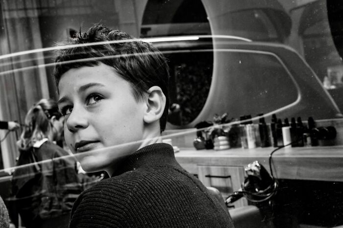 A young boy looking over his shoulder, with reflections in a window, capturing an authentic moment in street photography.