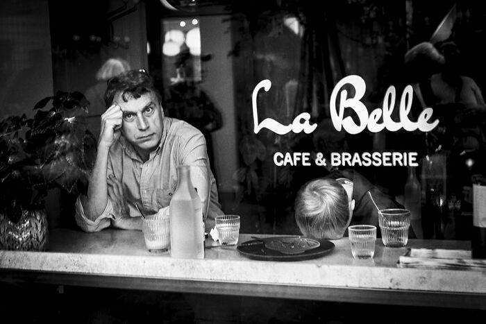 A man with his head in his hand looking at the camera, and another person with their head on the counter, in a café.