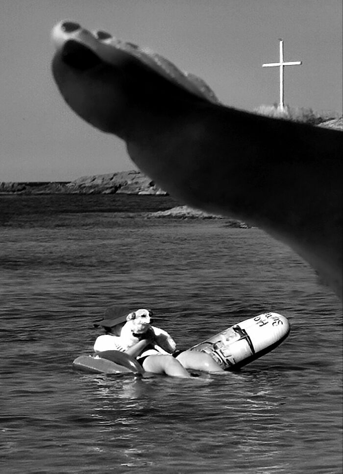 A black and white close-up of a foot, with a person floating in water and a cross in the background.