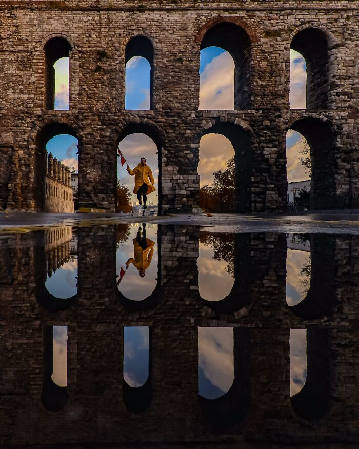 Murat Bakmaz captures the beauty of everyday life with a person reflected in a puddle before an arched stone wall.