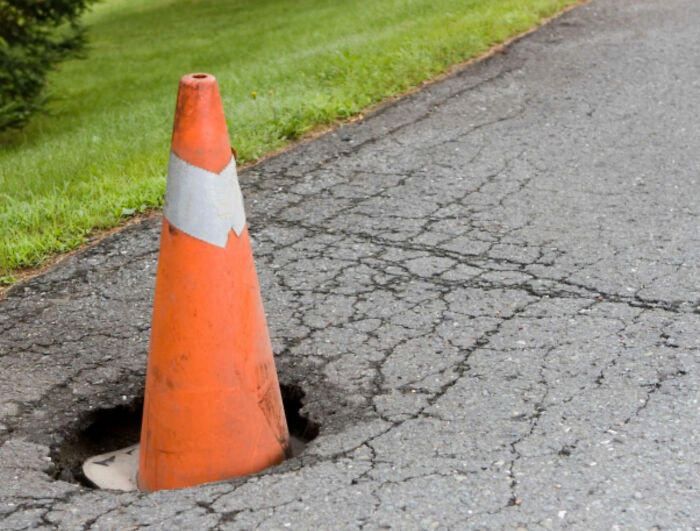 A vibrant orange traffic cone marking a prominent pothole on a cracked asphalt road in the USA.