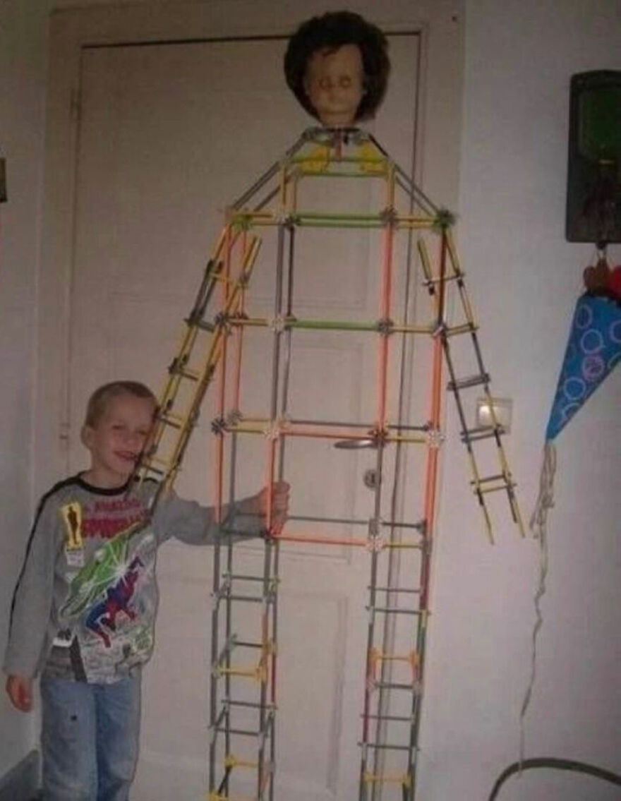 A smiling young boy standing next to a tall, human-like structure made of colorful sticks with a doll head, a random image.