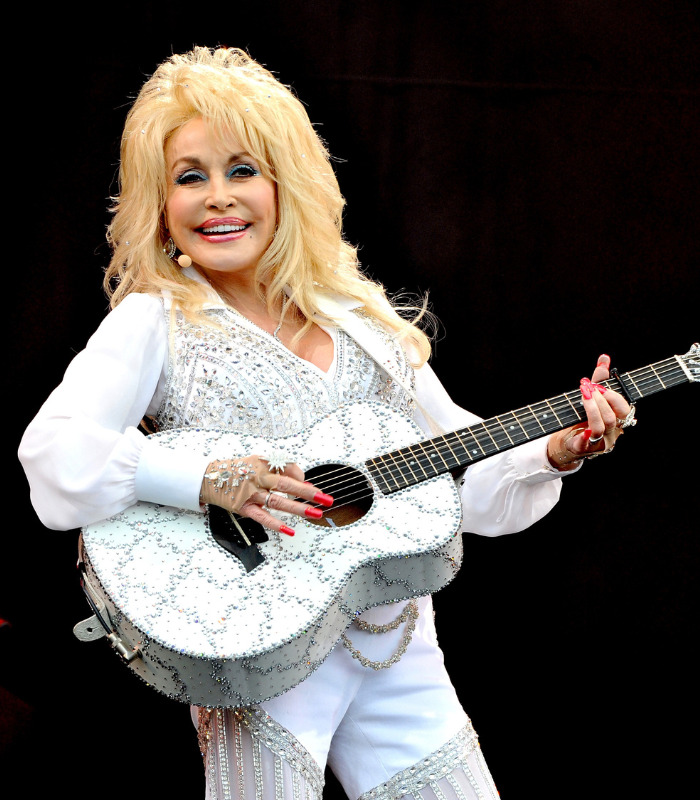 Country legend Dolly Parton performing on stage, playing a white, rhinestone-encrusted guitar. Country legend Dolly Parton performing on stage, playing a white, rhinestone-encrusted guitar.