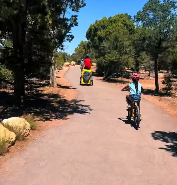 A kid on a bike and a parent on a recumbent bicycle exercising on a paved path through a sunny, tree-lined park. A kid on a bike and a parent on a recumbent bicycle exercising on a paved path through a sunny, tree-lined park.