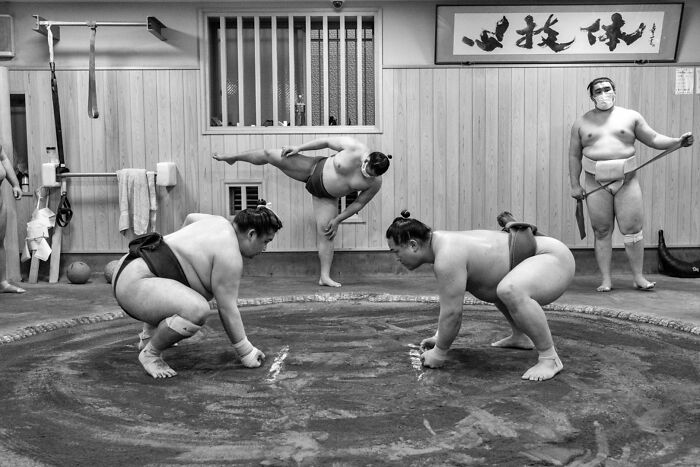 Black and white street photography scene showing three sumo wrestlers in a ring, one stretching, two squatting.