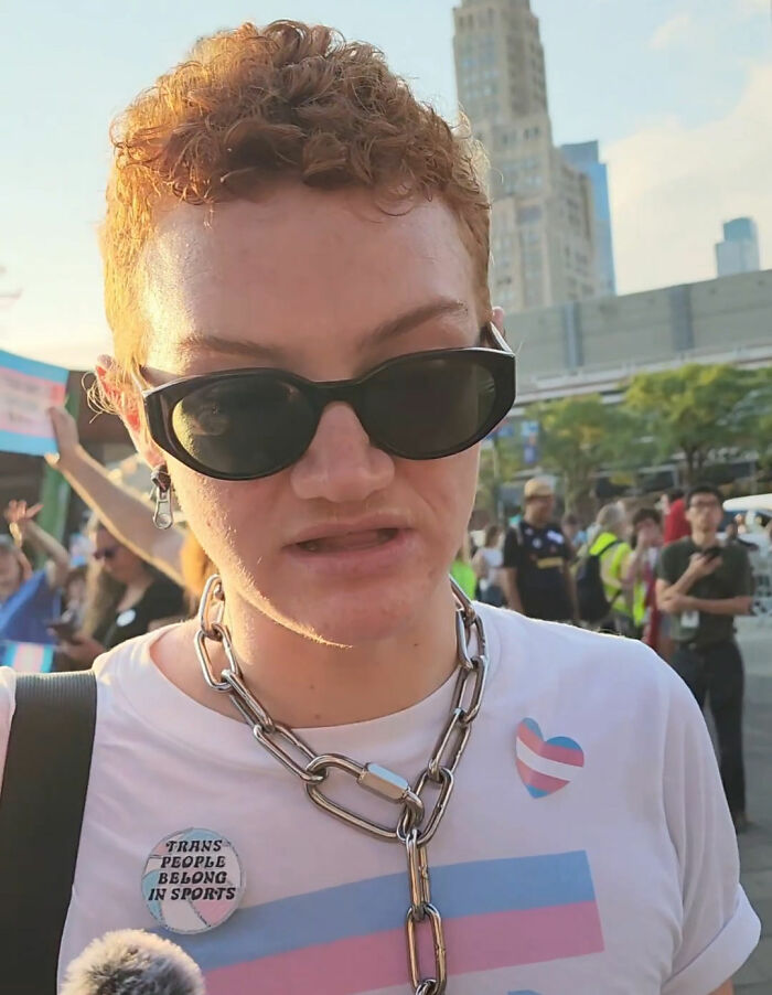 A person in sunglasses and a chain necklace at a protest, highlighting anti-trans protesters and WNBA question. A person in sunglasses and a chain necklace at a protest, highlighting anti-trans protesters and WNBA question.
