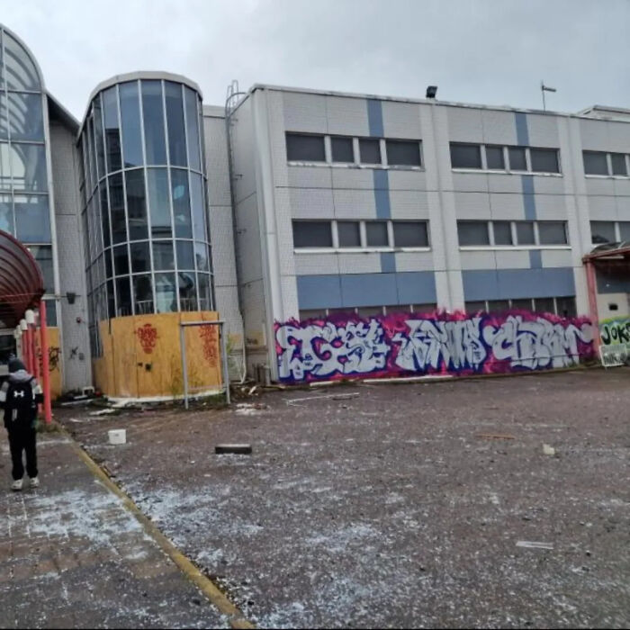 Urban exploration of an abandoned modern building with a glass facade, graffiti, and a debris-strewn ground.