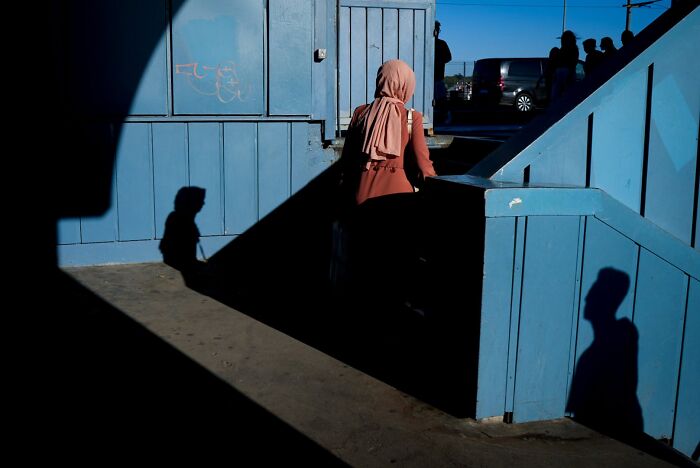 A woman in a hijab walks down blue stairs with stark shadows, a powerful street photography composition.