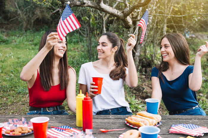 Three young women holding American flags at an outdoor picnic, enjoying American culture.