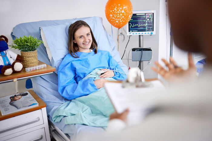 A smiling woman in a hospital bed with a get well balloon, relating to absurd American questions to Europeans.