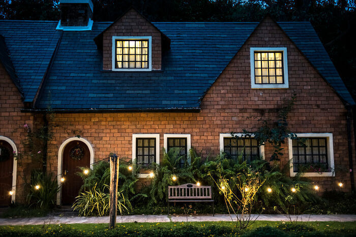 Charming brick house with glowing windows and string lights, reminiscent of a cozy European home, contrasting with absurd American questions.