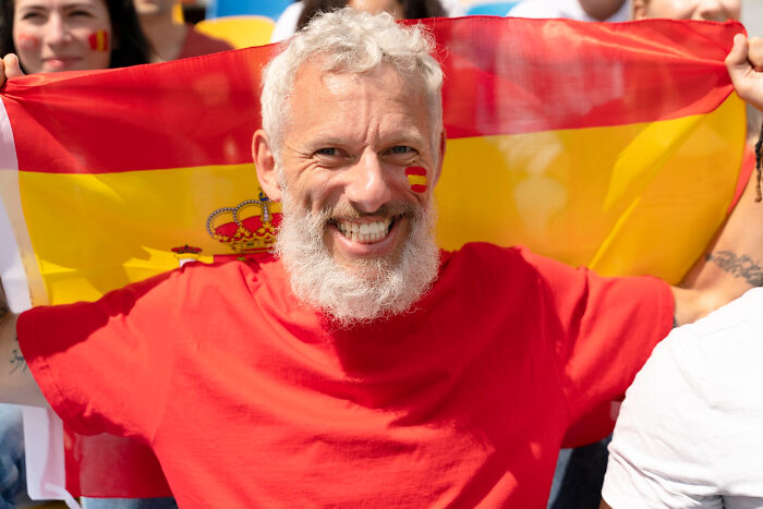 A smiling man with a beard holds a Spanish flag, representing Europeans. He has a Spanish flag painted on his cheek, ready for absurd American questions.