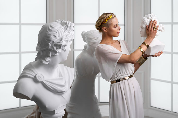 Absurd American questions: a woman in a white dress holding a bust sculpture, surrounded by other statues.