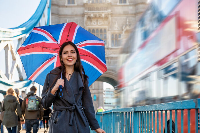 Woman with a Union Jack umbrella in London, featuring Tower Bridge and a red bus, representing European culture and absurd American questions.