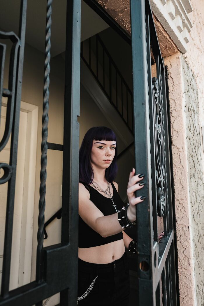 A young woman with purple hair and dark clothing standing behind a metal gate, showing a person seeing red.