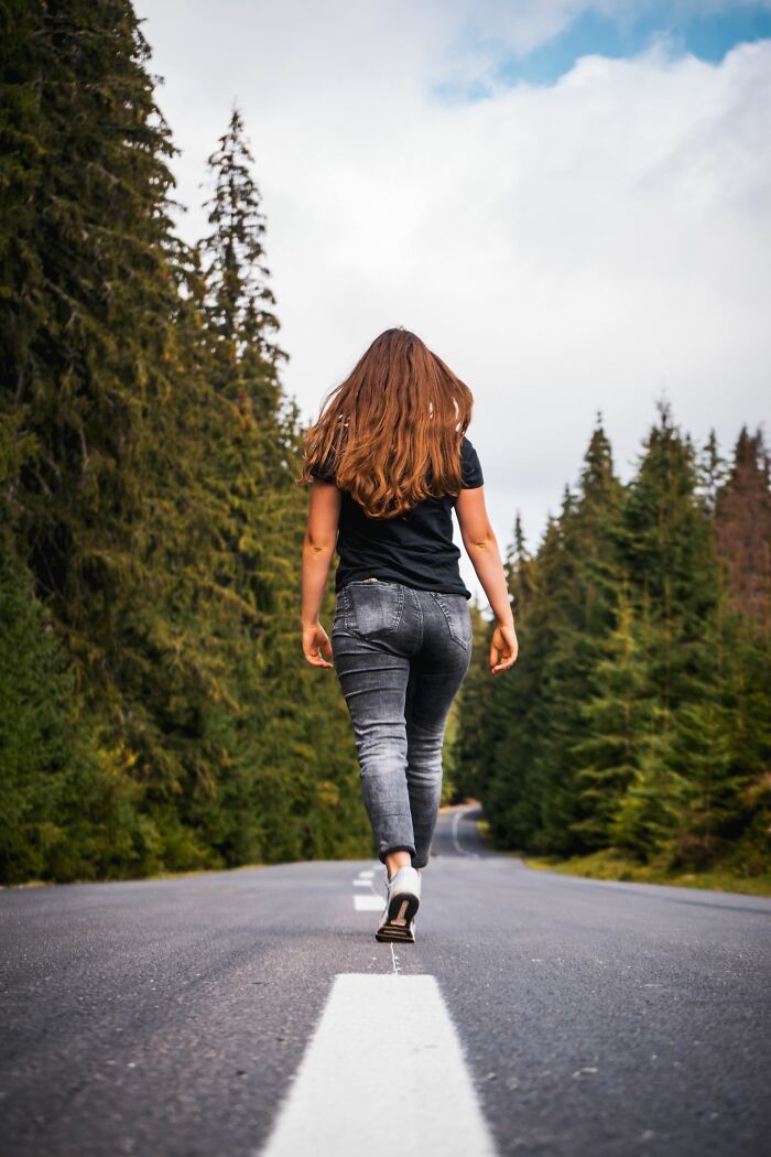 A woman with long brown hair walking away on a road surrounded by tall trees, representing ridiculous reasons.