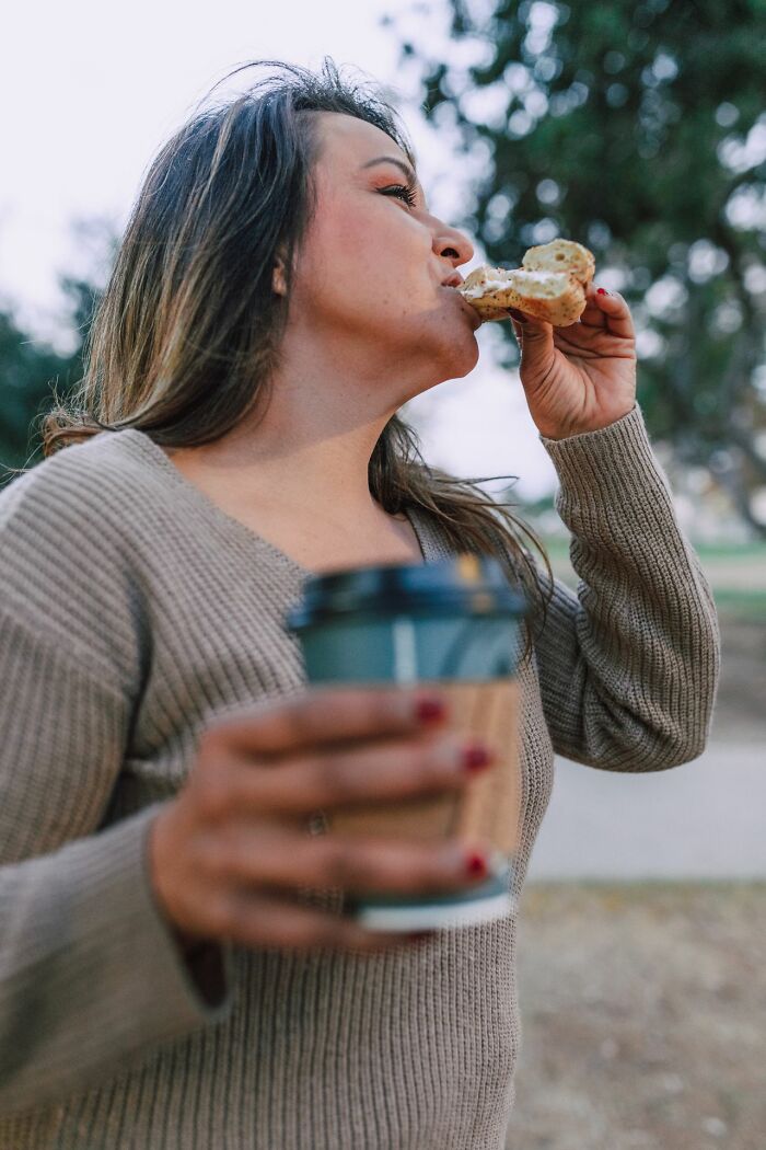 A woman eating a bagel and holding coffee, ignoring someone due to ridiculous reasons for not speaking.