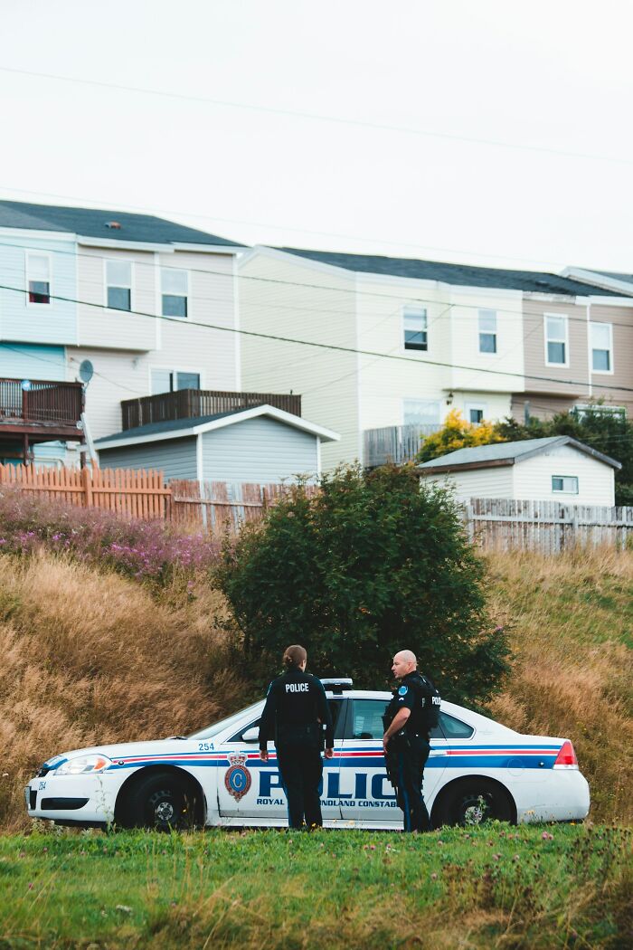 Two police officers stand next to a patrol car on a grassy hill, with houses in the background. This image shows ridiculous reasons people stop speaking to each other.