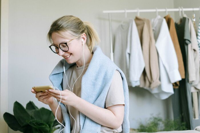 A smiling woman wearing glasses and earphones looks at her phone. This image represents ridiculous reasons people stop speaking to each other.