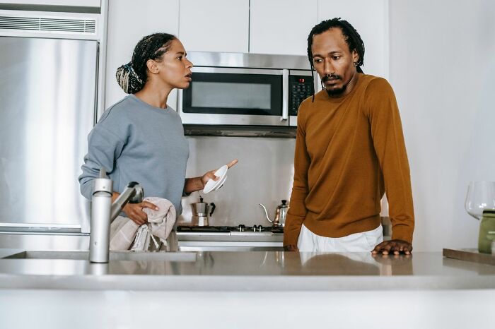 A couple in a kitchen, a woman upset, showing ridiculous reasons for not speaking.