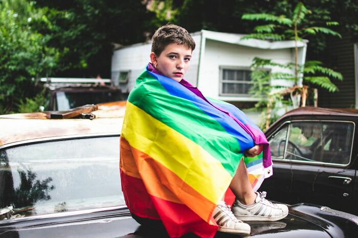 A person with short hair wrapped in a rainbow flag sits on the hood of an old car. This image represents ridiculous reasons people stop speaking to each other.
