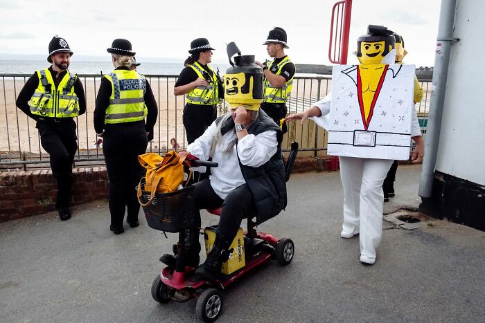 People in LEGO block costumes, one on a scooter and one dressed as Elvis, interacting with police officers, a humorous street photography moment.
