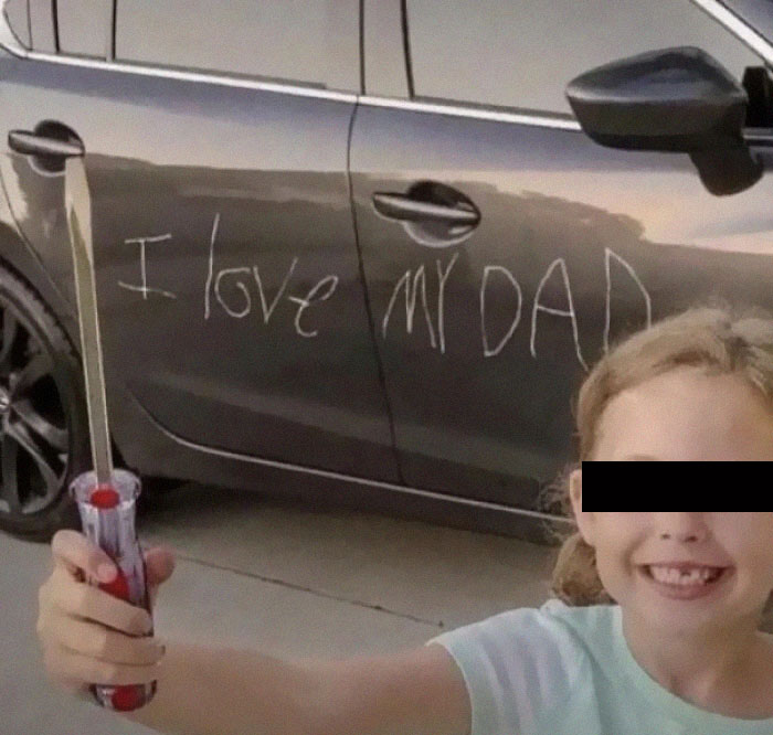 A child smiling while holding a screwdriver next to a car with 'I love my dad' scratched into it, a memorable moment for parents.