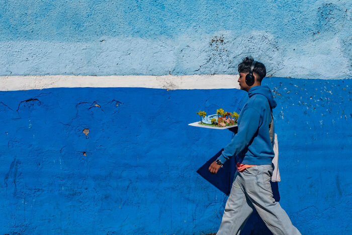A man in headphones walks past a blue wall carrying a tray, an artistic street photography award scene.