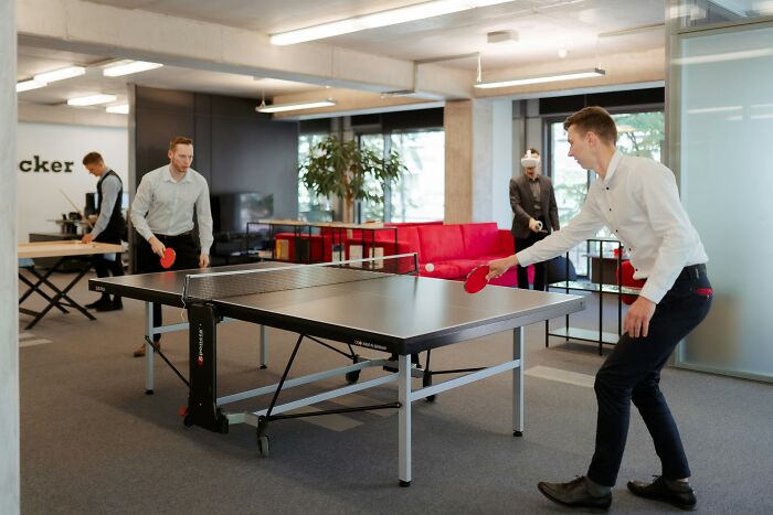 Two men playing table tennis in an open-plan office, highlighting work drama stories during boring office hours.