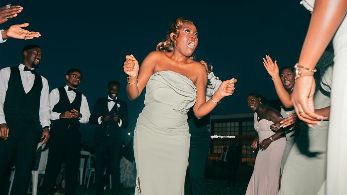 A group of guests dancing enthusiastically at a wedding reception, featuring a woman in a sage green dress.