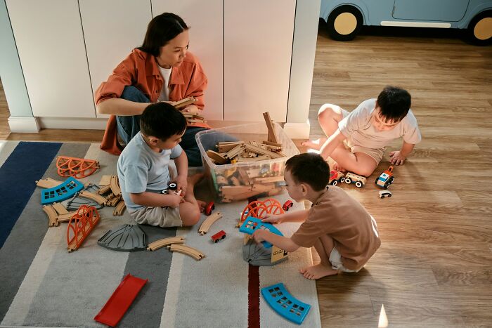 A woman and three young children play with wooden toy trains on a rug. Horrible parent.