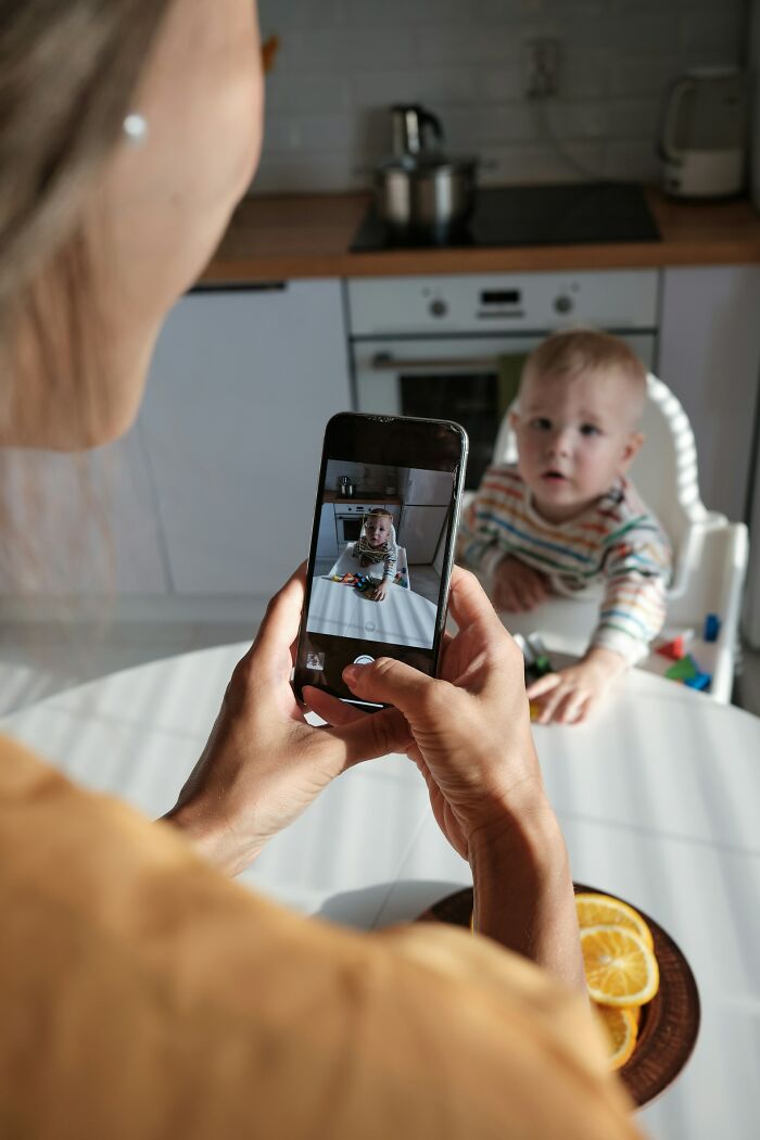 A parent taking a picture of a baby in a highchair with a smartphone. This represents a parent capturing a moment, but sometimes parenting is horrible.