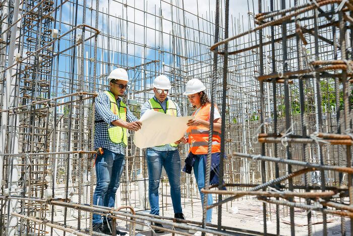 Three construction workers in hard hats and safety vests examining blueprints at a building site with exposed rebar, showcasing industry secrets.