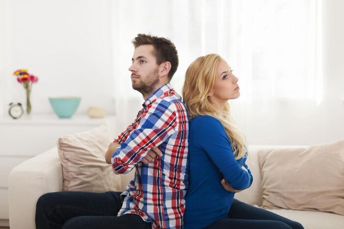 A couple sitting back-to-back on a sofa, arms crossed, both looking away from each other, illustrating unhinged house rules in a family relationship.