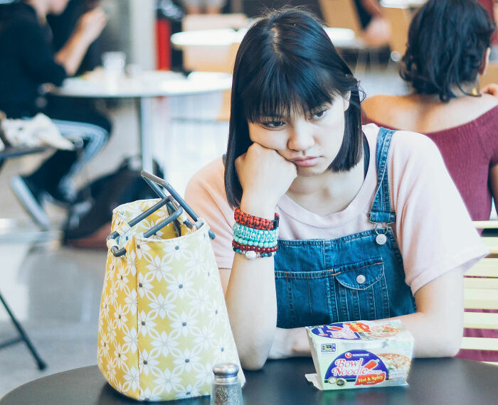 A young woman looking sad while sitting at a table with a package of instant noodles and a lunch bag, comparing school lunch in France to USA. A young woman looking sad while sitting at a table with a package of instant noodles and a lunch bag, comparing school lunch in France to USA.