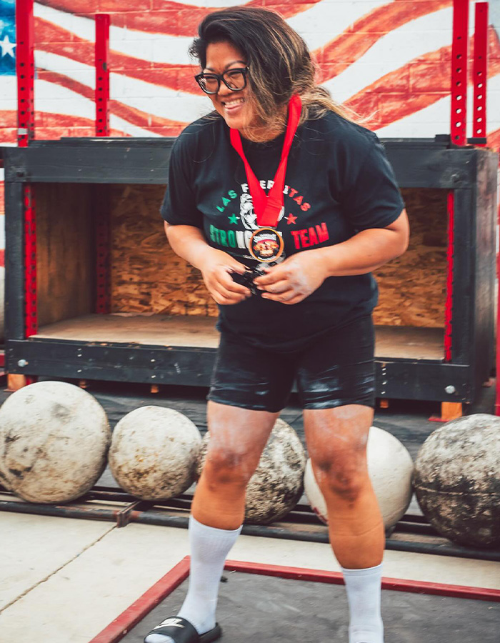 A strength athlete with a medal around her neck, smiling broadly, standing near large stone weights.