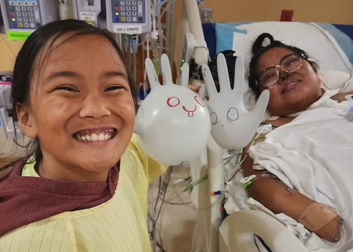 A young girl smiling and holding up two white glove balloons for a strength athlete in a hospital bed.
