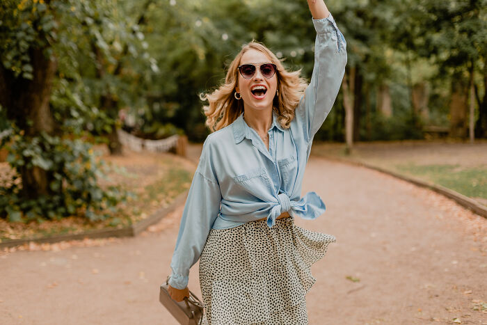 Woman in sunglasses and a denim shirt cheering outdoors, a real deal sign from the universe.