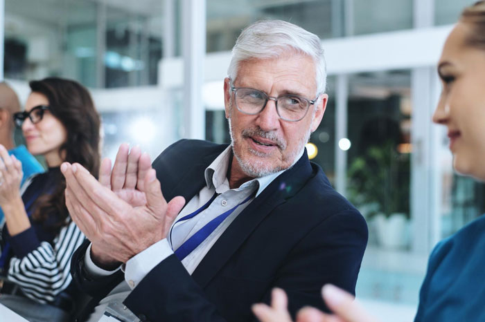 A man with grey hair and glasses claps, engaged in a charity auction event. A man with grey hair and glasses claps, engaged in a charity auction event.