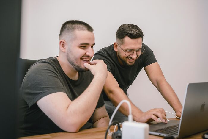 Two men smiling while looking at a laptop, reading work drama stories.